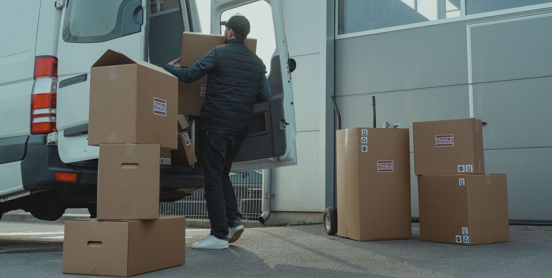 A delivery man unloading cardboard boxes from a van at a warehouse during the day.