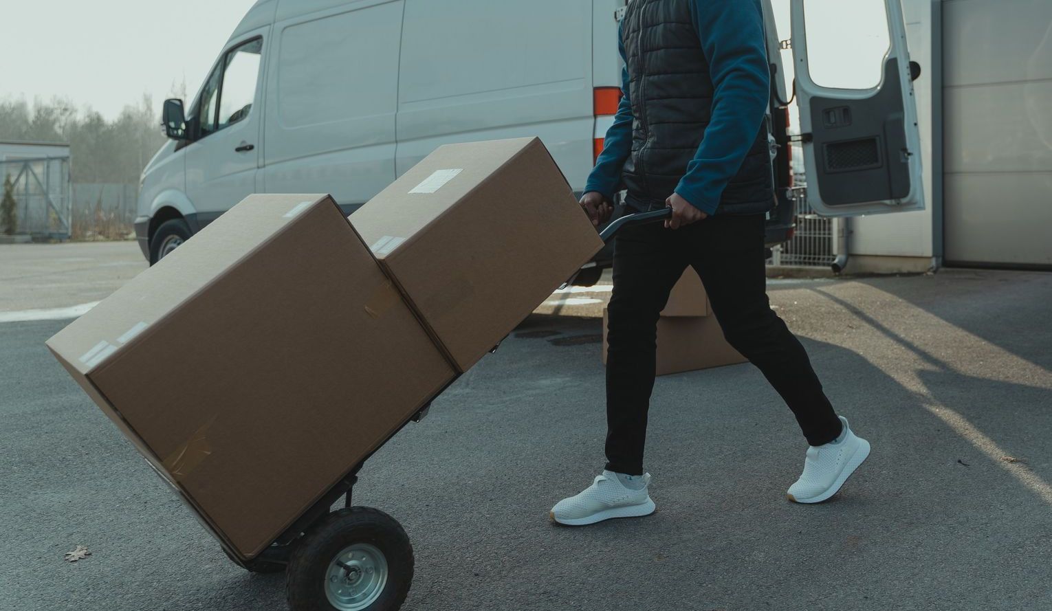 Courier pushing a trolley with boxes near a van at a warehouse, showcasing delivery services.